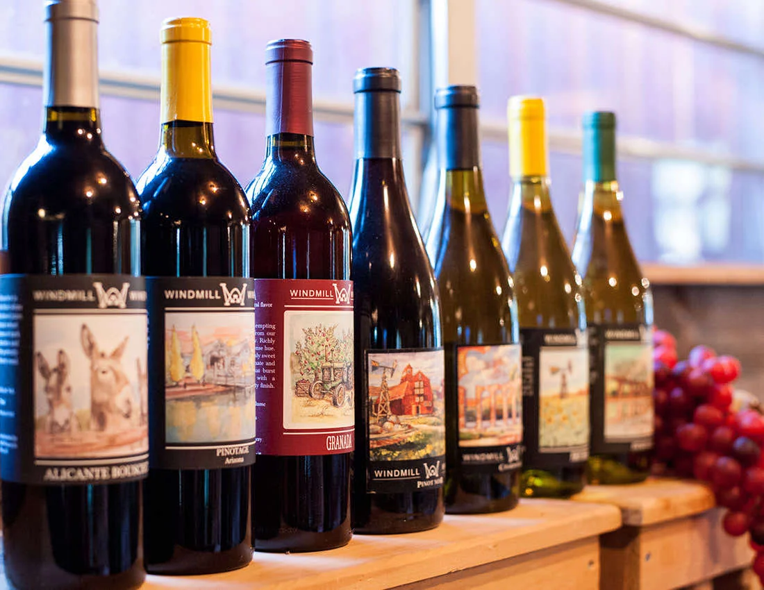 A lineup of Windmill wine bottles with illustrated labels, beside a cluster of red grapes